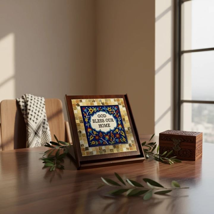 Handmade ceramic and mosaic plaque featuring “God Bless Our Home” in the center, surrounded by colorful Palestinian patterns, olive branches, and a carved wooden box, displayed on a wooden table in natural light